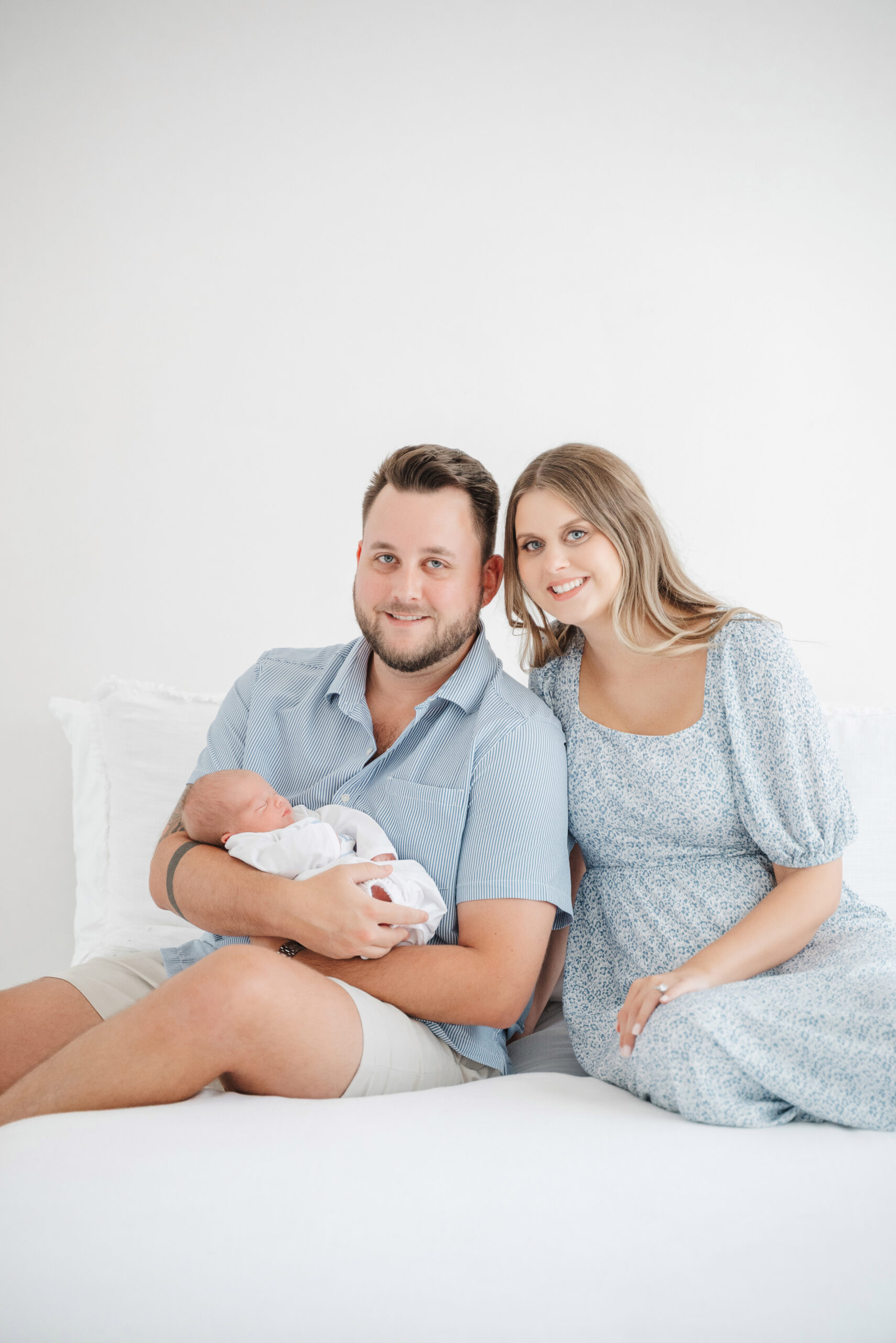 Mom and dad snuggle close on bed while holding newborn baby boy in Cumming Newborn photography session.