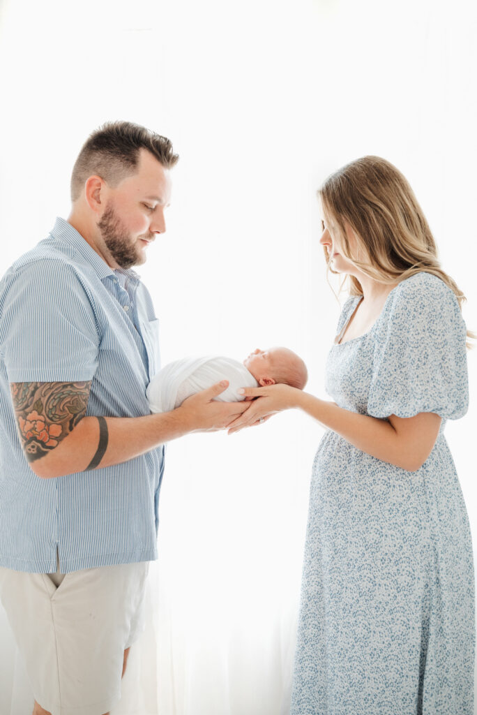 Mom and dad stand in front of a window and look down a their newborn baby inin Cumming newborn photoshoot.