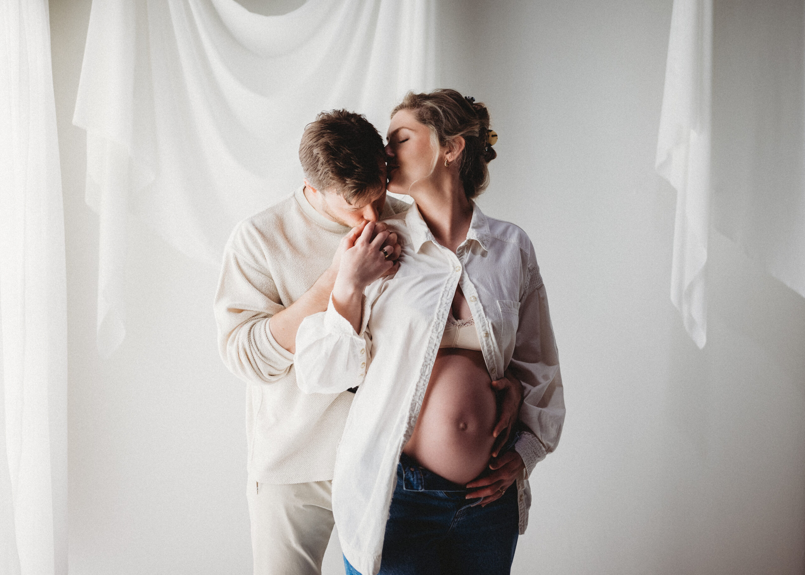 Husband embraces wife and kisses her hand as she poses in white open button down showing pregnant belly