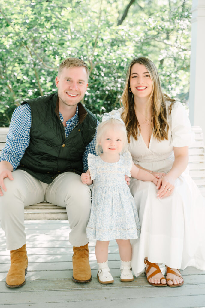 Family of three posing together on a porch bench in Alpharetta GA: mom, dad, and toddler daughter smiling outdoors