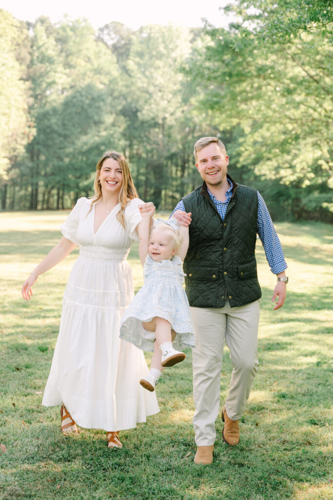 Parents swinging toddler daughter between them while walking outdoors in Alpharetta GA. Happy family enjoying time together outside
