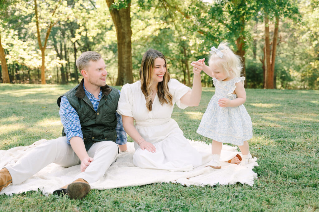 Happy family sitting outdoors in Alpharetta Georgia: dad, mom, and toddler daughter laughing together in a sunny park setting