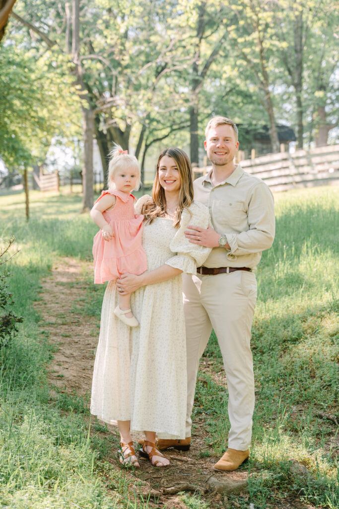 Family of three standing on a sunny outdoor path in Alpharetta GA:  mom holding toddler daughter in a pink dress while dad smiles beside them