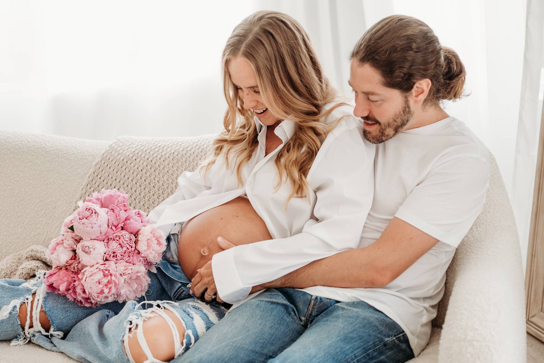 A pregnant couple snuggles on a couch in white shirts and jeans while holding the bump