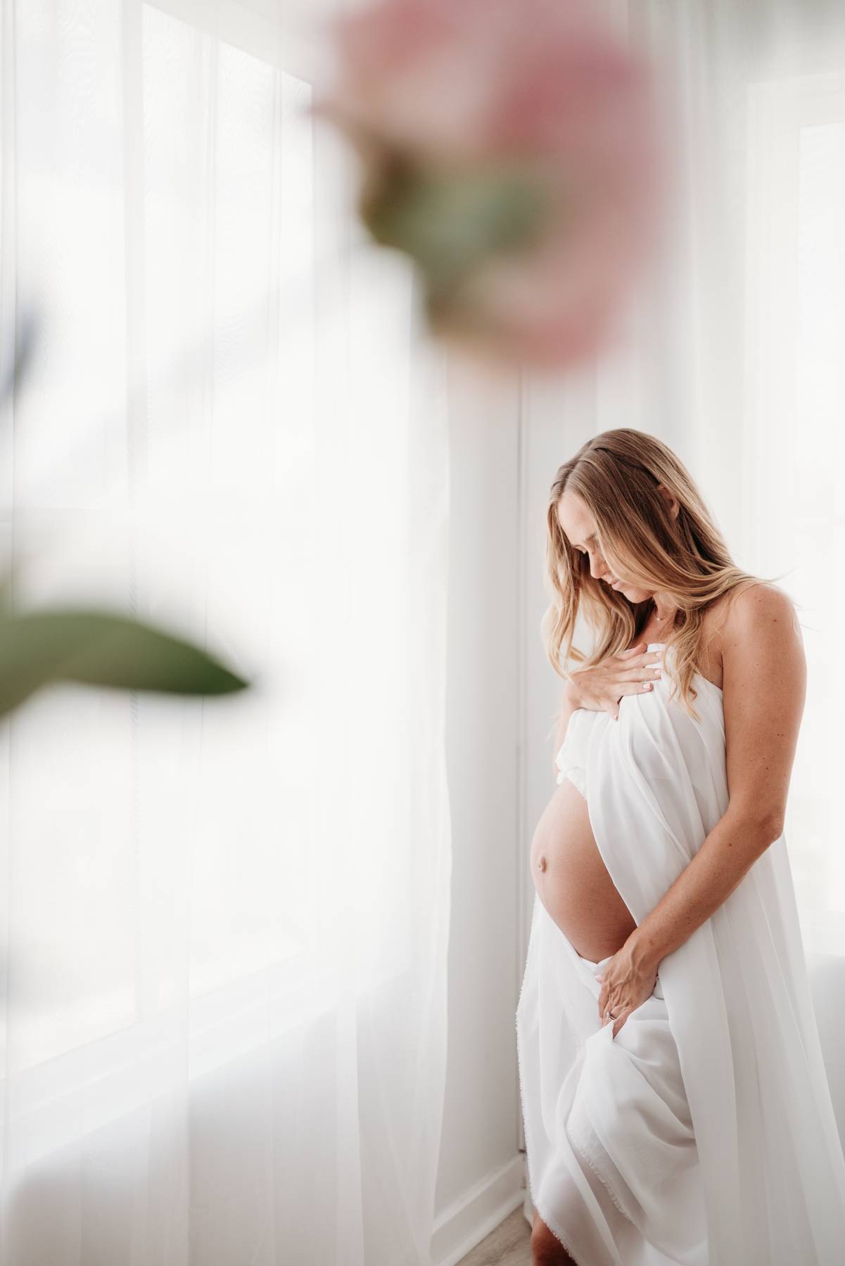 A mom to be wrapped in a white sheet gazes down at her bump while standing in a window after a 3d ultrasound in atlanta