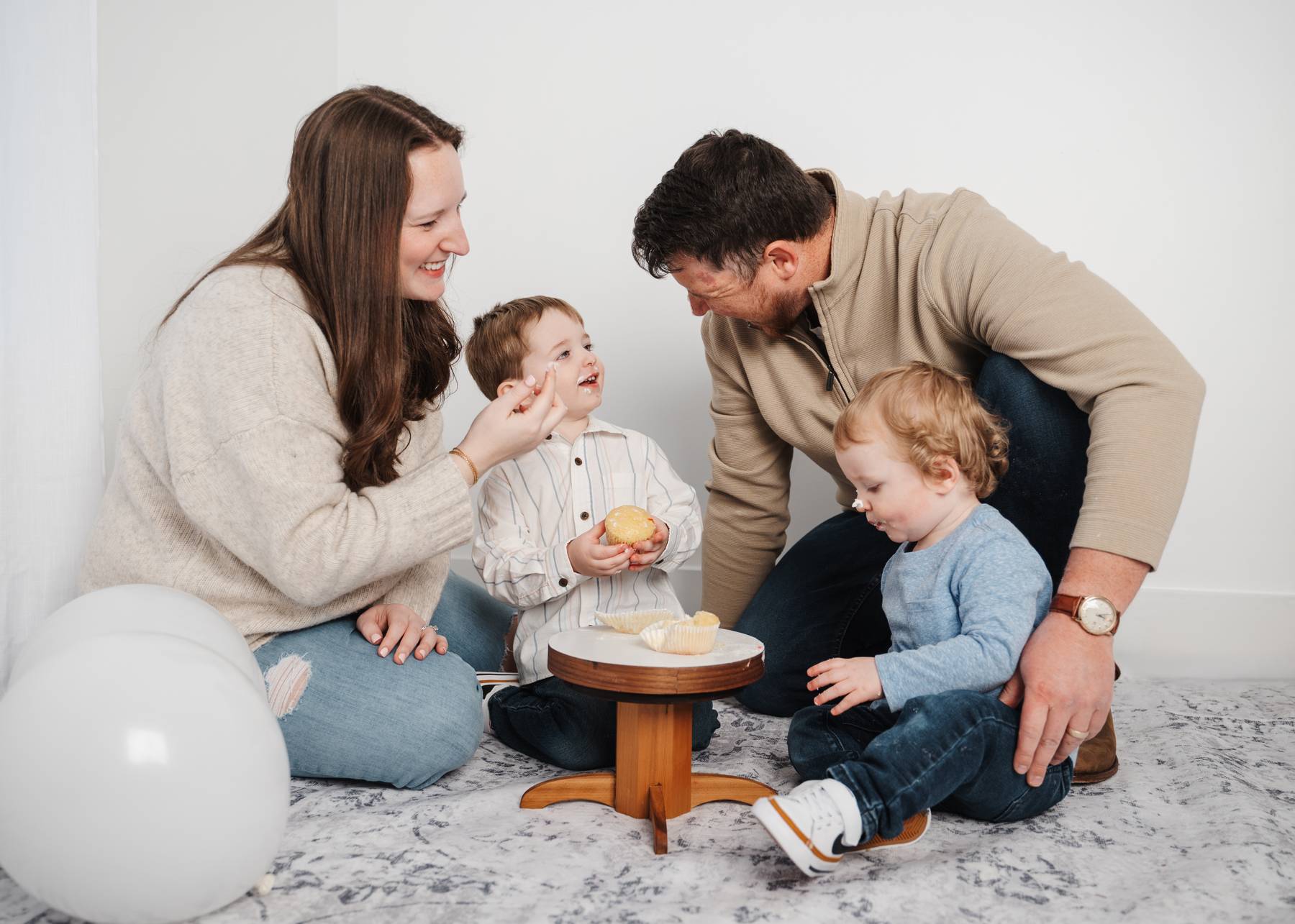 Toddler brothers eat cupcakes with laughing mom and dad on a rug in a studio after visiting alpharetta daycares