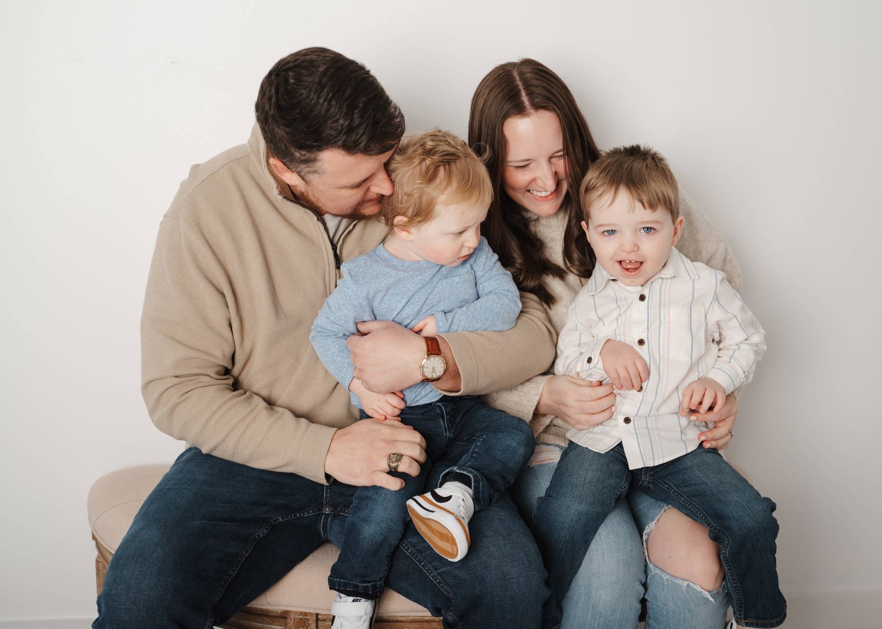 Giggling mom and dad sit on a bench in a studio tickling their toddler sons in their laps after visiting alpharetta daycares