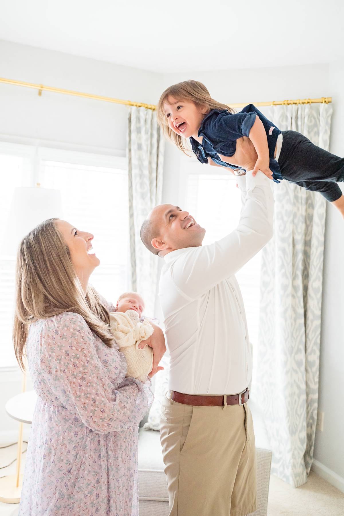 An ecstatic toddler girl laughs while dad lifts her above his head as mom holds a newborn before some gymnastics in Buford, GA