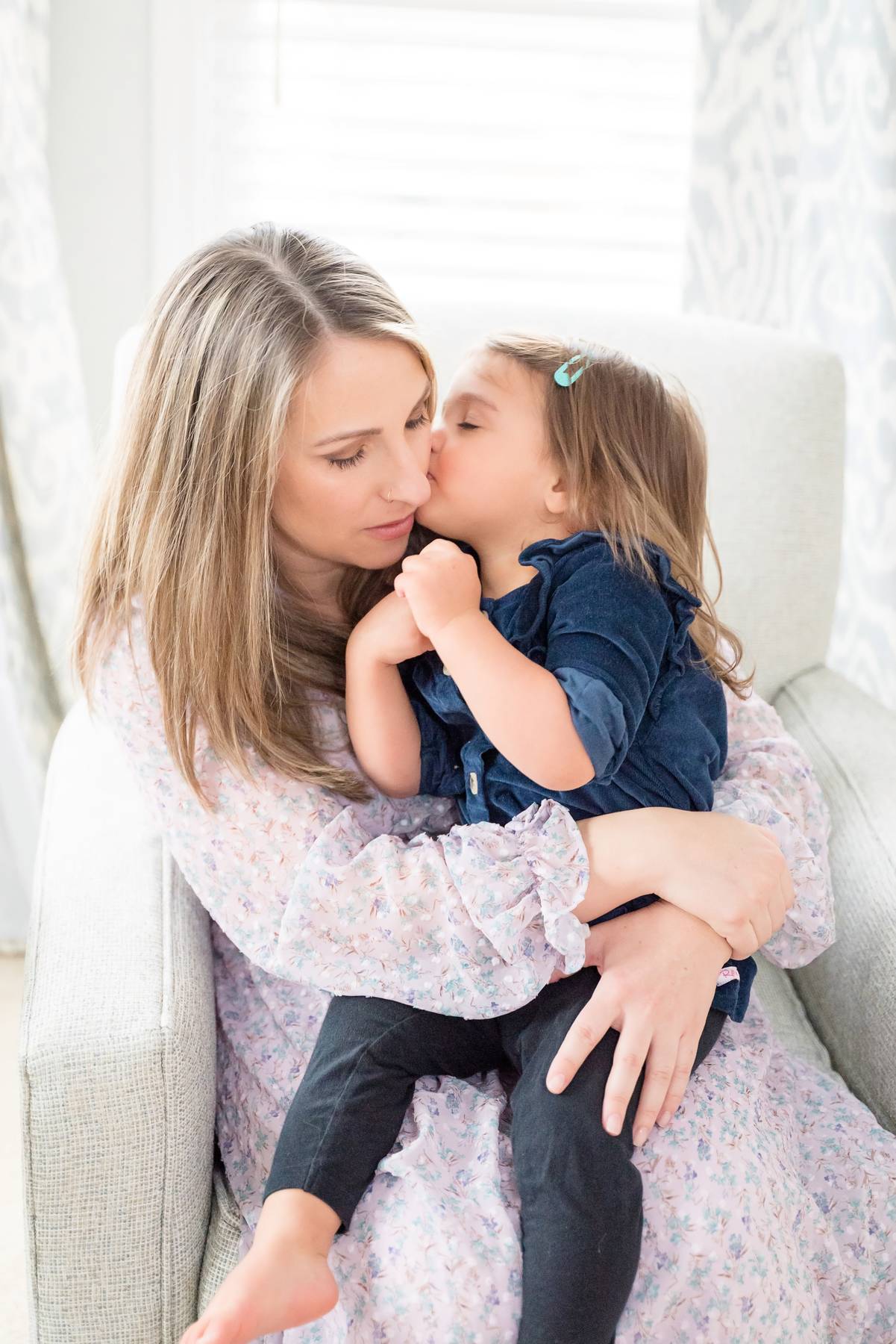 A toddler girl in blue whispers in mom's ear while sitting in her lap in a chair before some gymnastics in Buford, GA