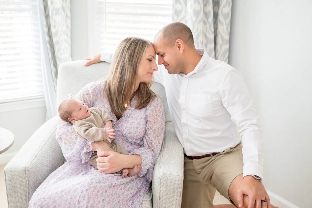 Happy new parents touch foreheads while mom sits in a nursing chair holding their baby