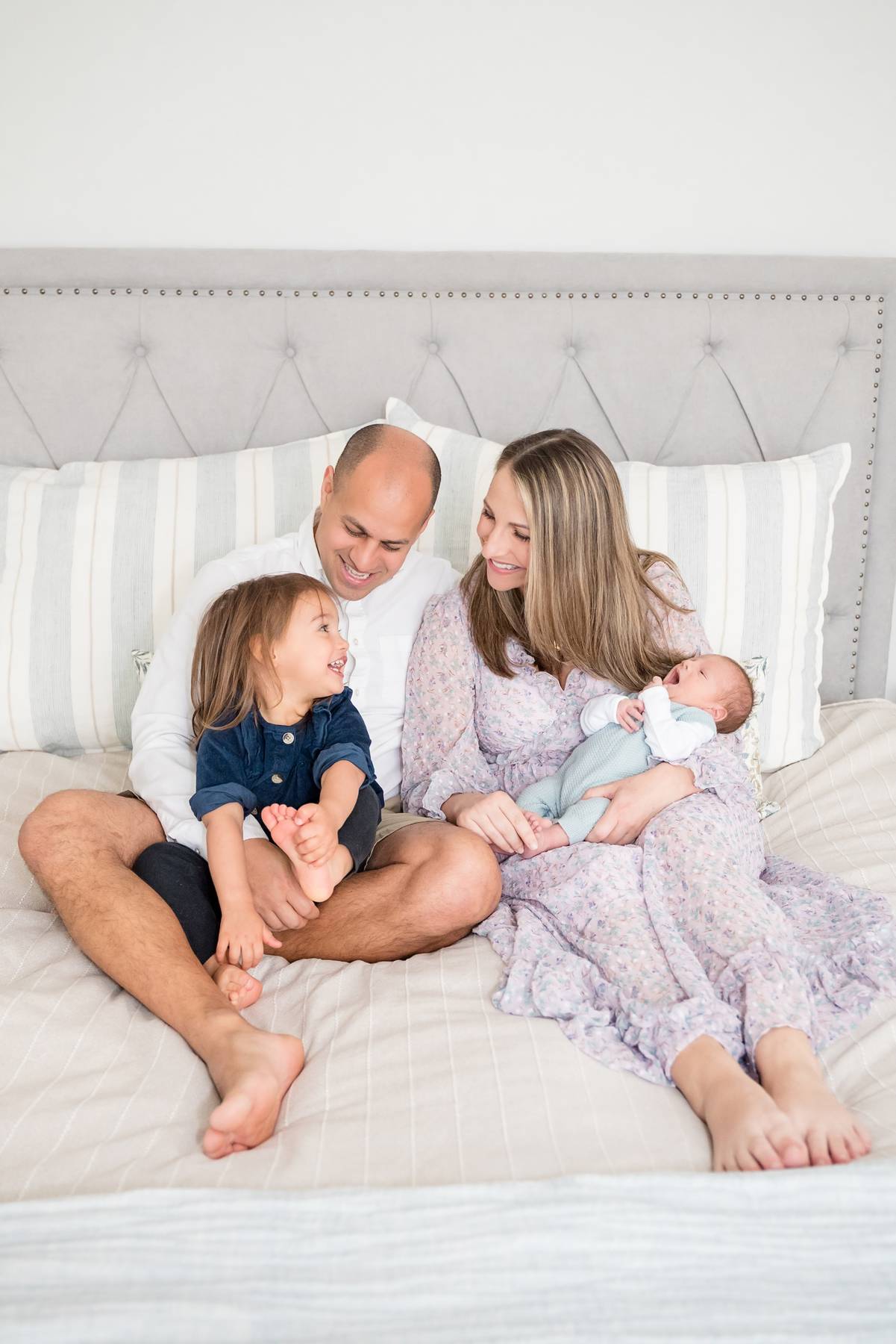A happy family sits on a bed playing with a toddler and newborn baby in mom and dad's laps after using an obgyn in alpharetta ga