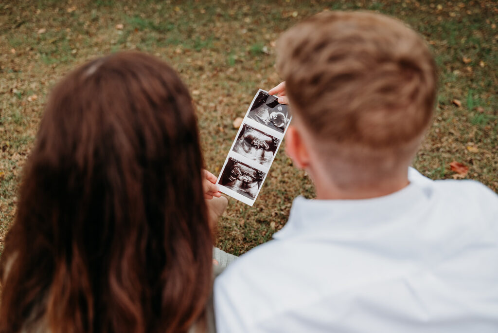Couple holding an ultrasound photo while sitting in autumn leaves.