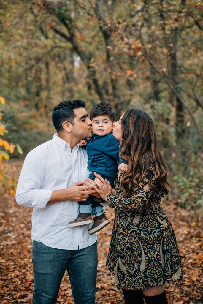 Parents kissing toddler’s cheeks during Cumming GA fall family portraits in autumn leaves