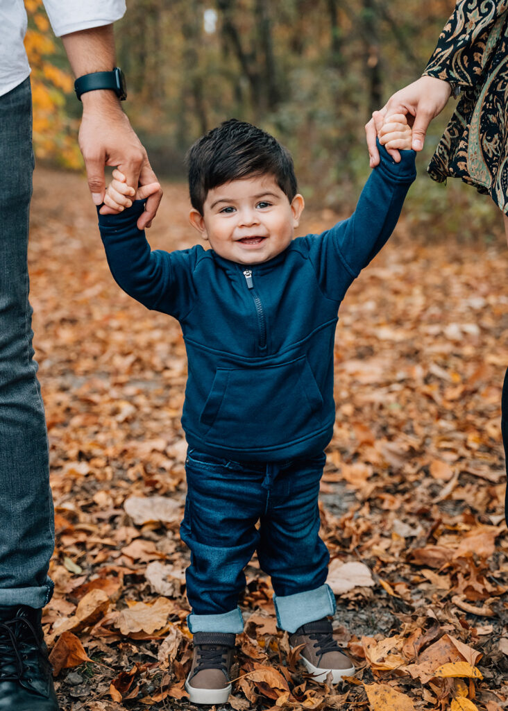 Toddler holding parents’ hands while walking through fall leaves in Cumming GA family portraits