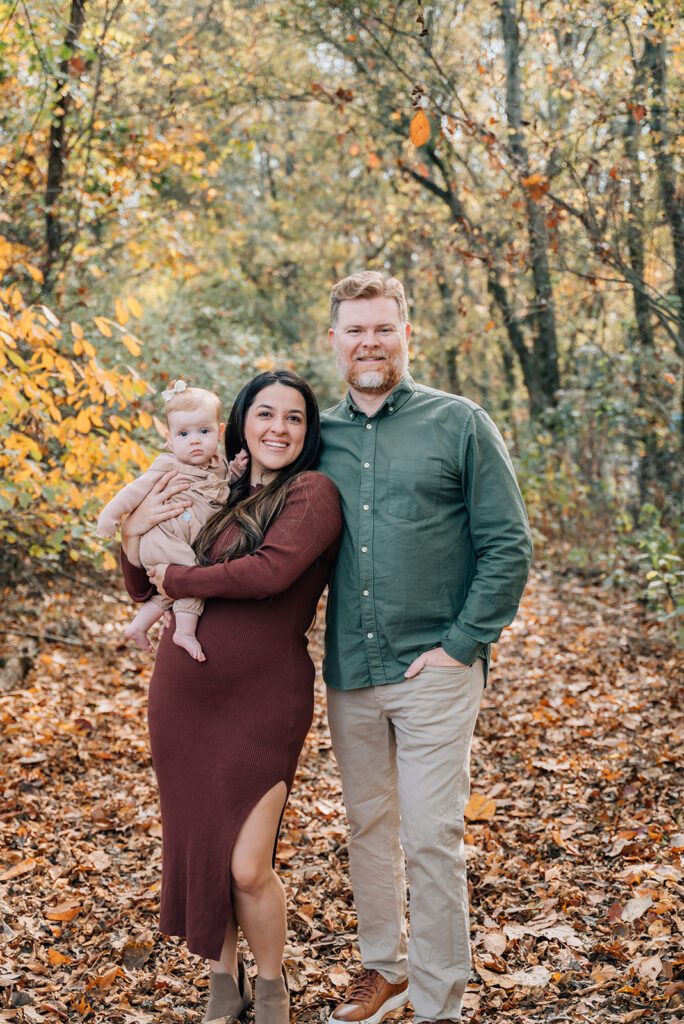 Young family smiling during fall family photo session in North Georgia woods, mom holding baby girl in beige romper, dad in green button-down shirt