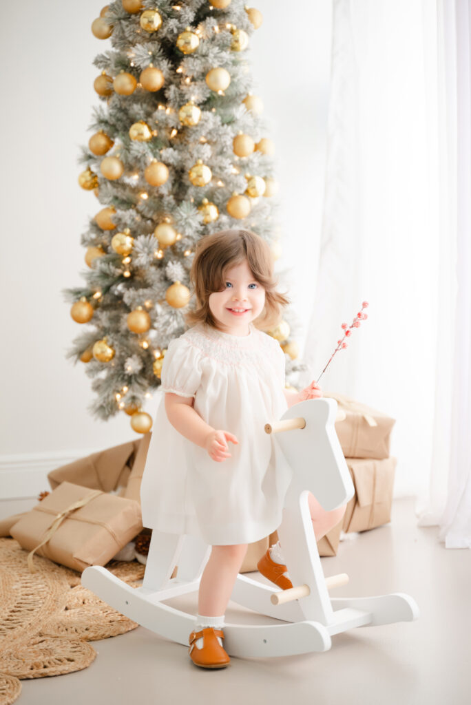 Little girl on rocking horse wearing a white christmas outfit for family photos.