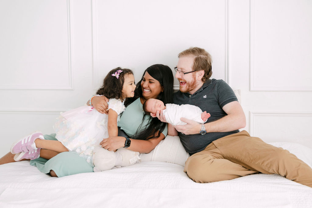 Mom, daughter, dad and baby sit together and laugh on white daybed. 