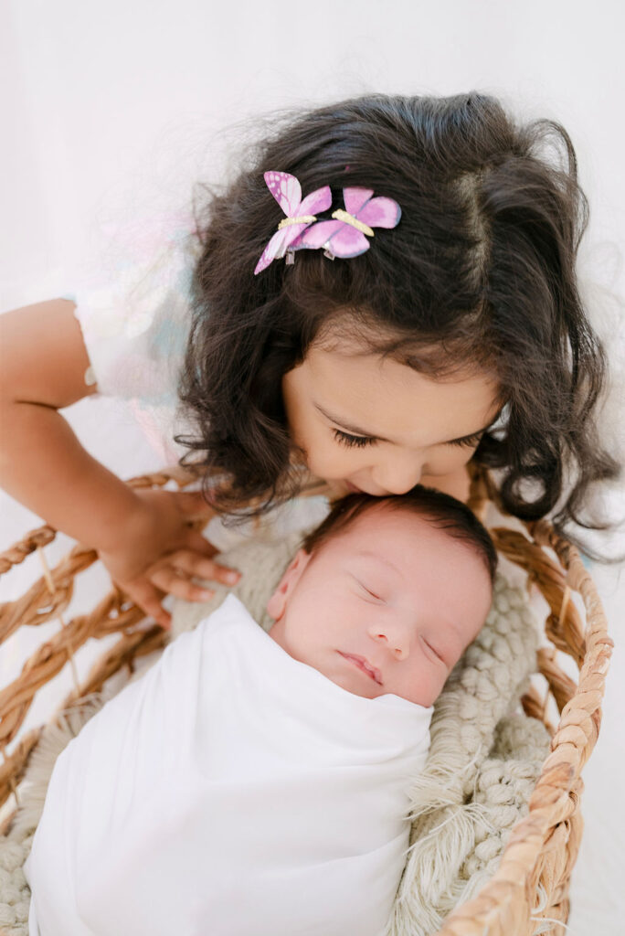 Girl kisses newborn brother who is swaddled in a basket in a newborn session in Alpharetta.