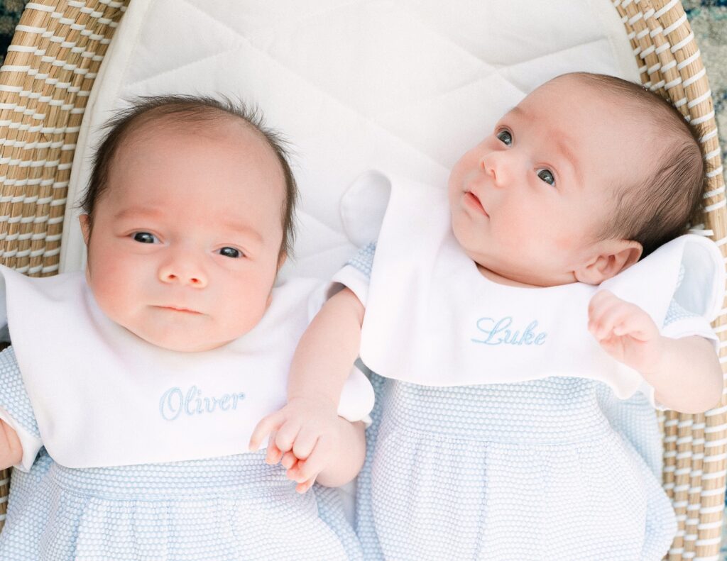 Twin newborn boys in matching blue outfits hold hands while laying in a bassinet in Atlanta Newborn Photography session. 