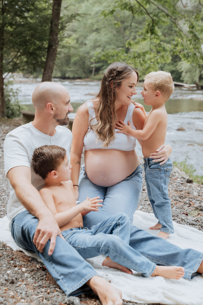 Pregnant mom sits with dad, and two sons in front of river.