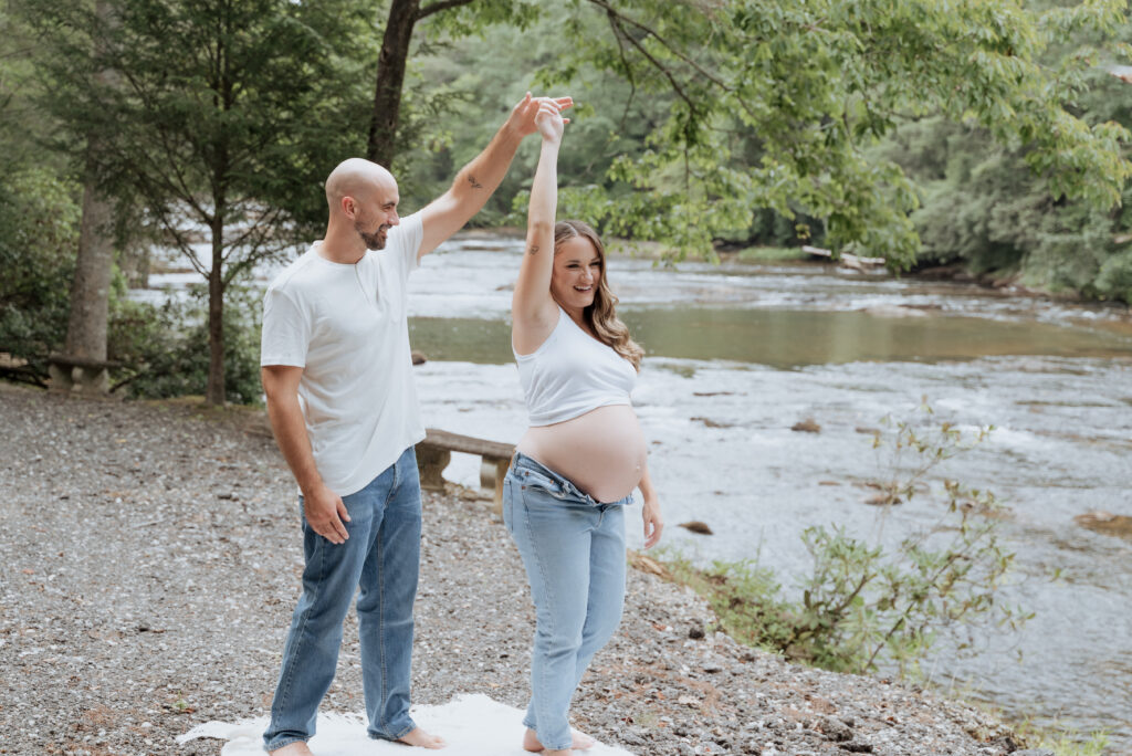 Husband dances with pregnant wife in front of river in maternity session.