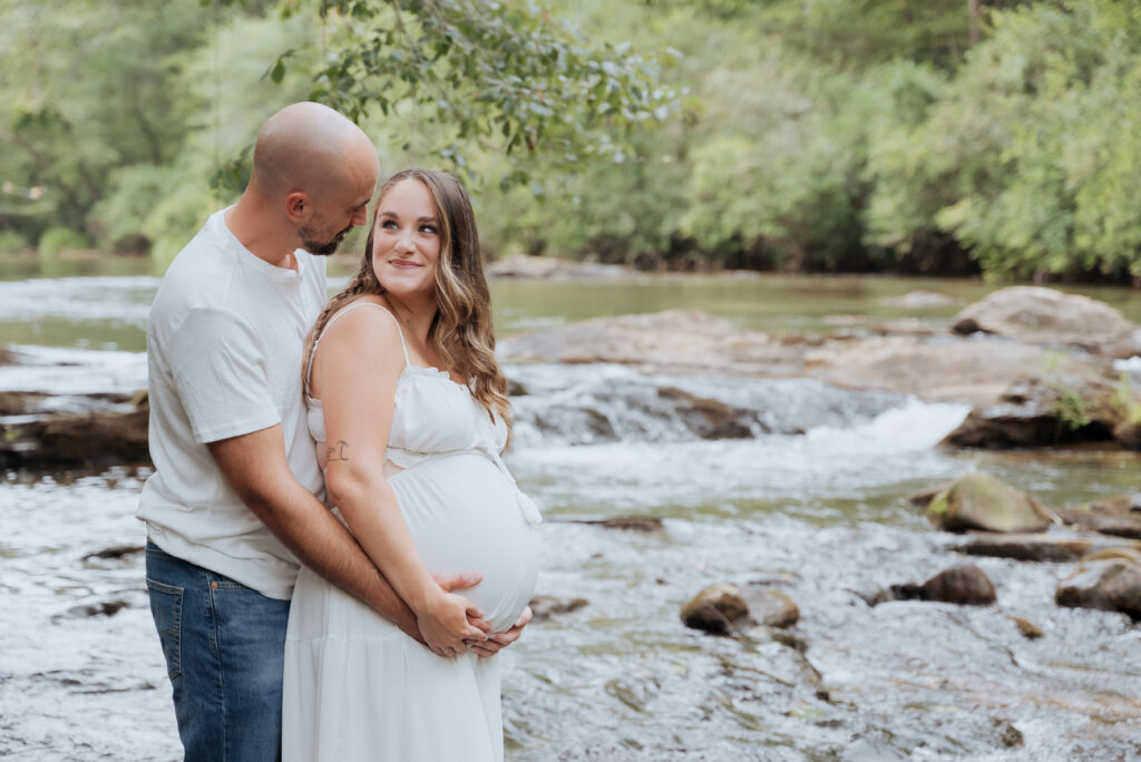 Mom and dad look into eachother's eyes while standing in river during Maternity photoshoot.