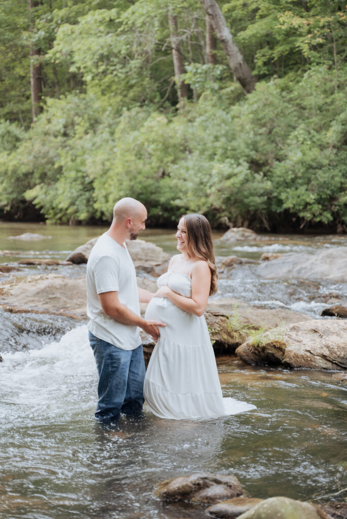 Mom and dad look into each others eyes while laughing and holding pregnant belly in river.