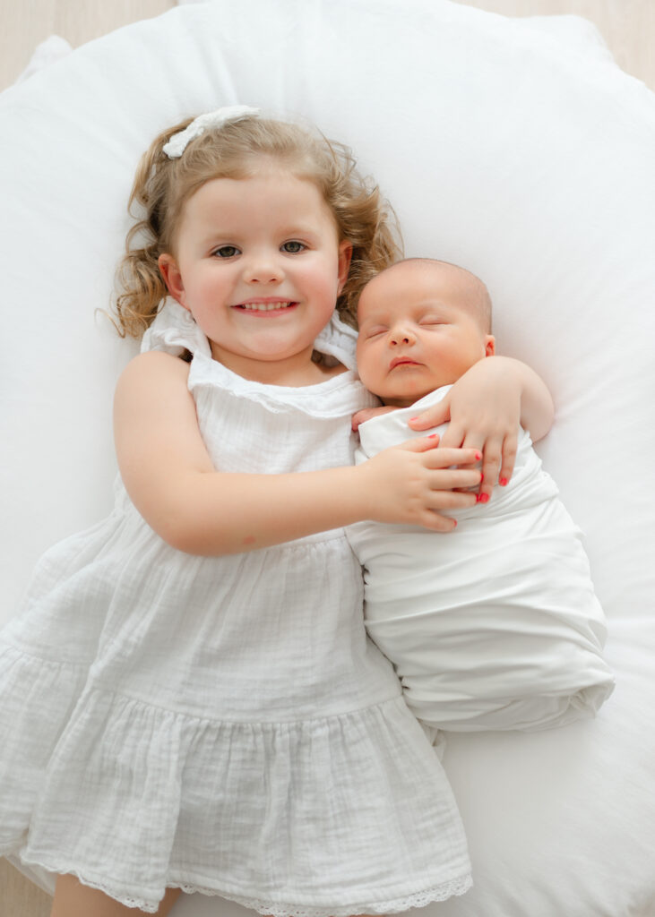 Big sister hugs little brother wrapped in white swaddle on white beanbag during Atlanta newborn photography session. 